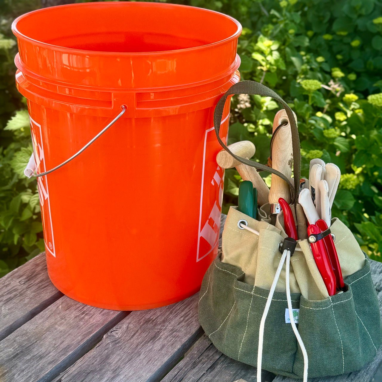Orange bucket and green tool pouch with gardening tools on a wooden surface with greenery in the background