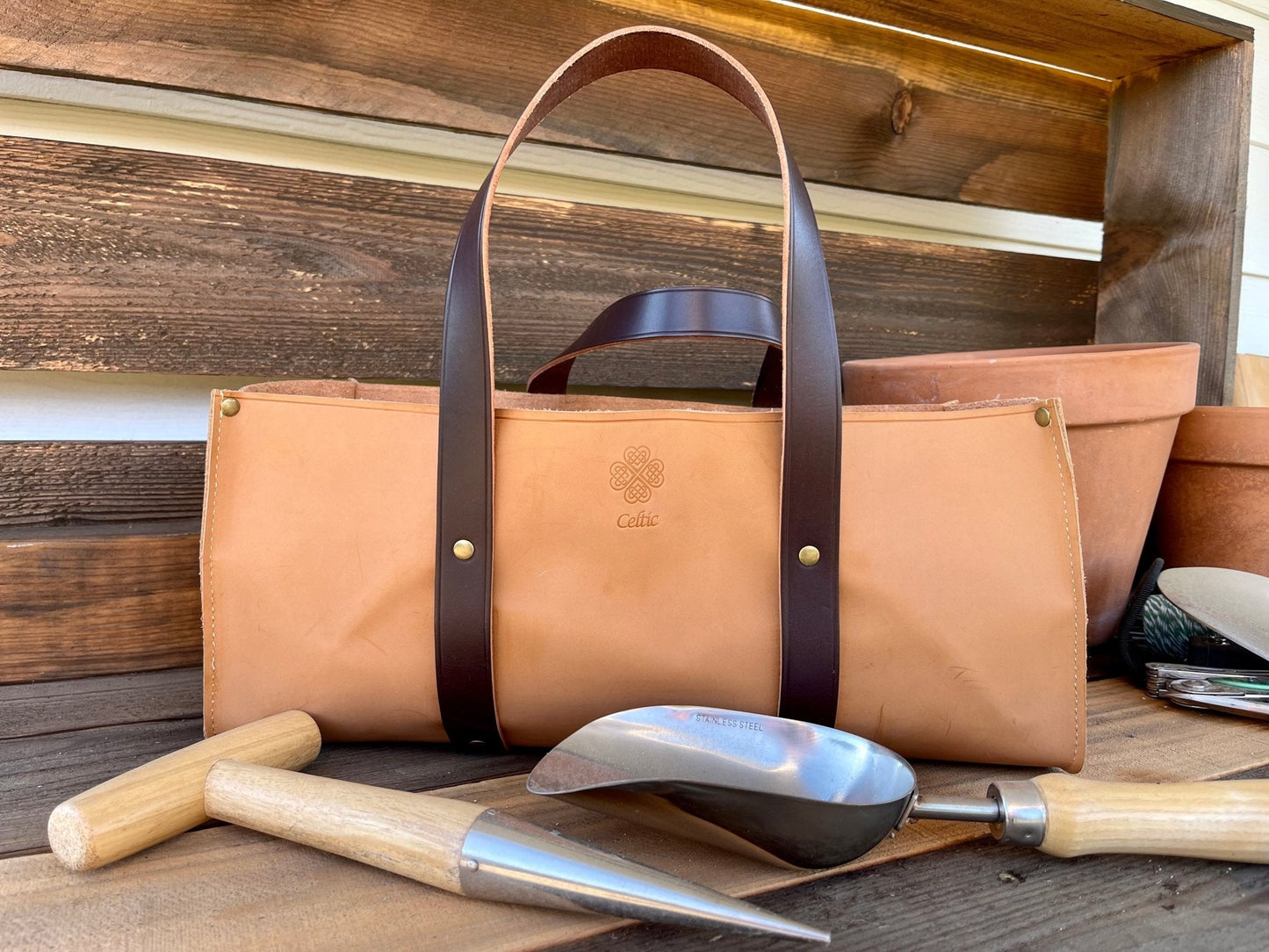 Brown leather tote bag with handles on a wooden surface with gardening tools.