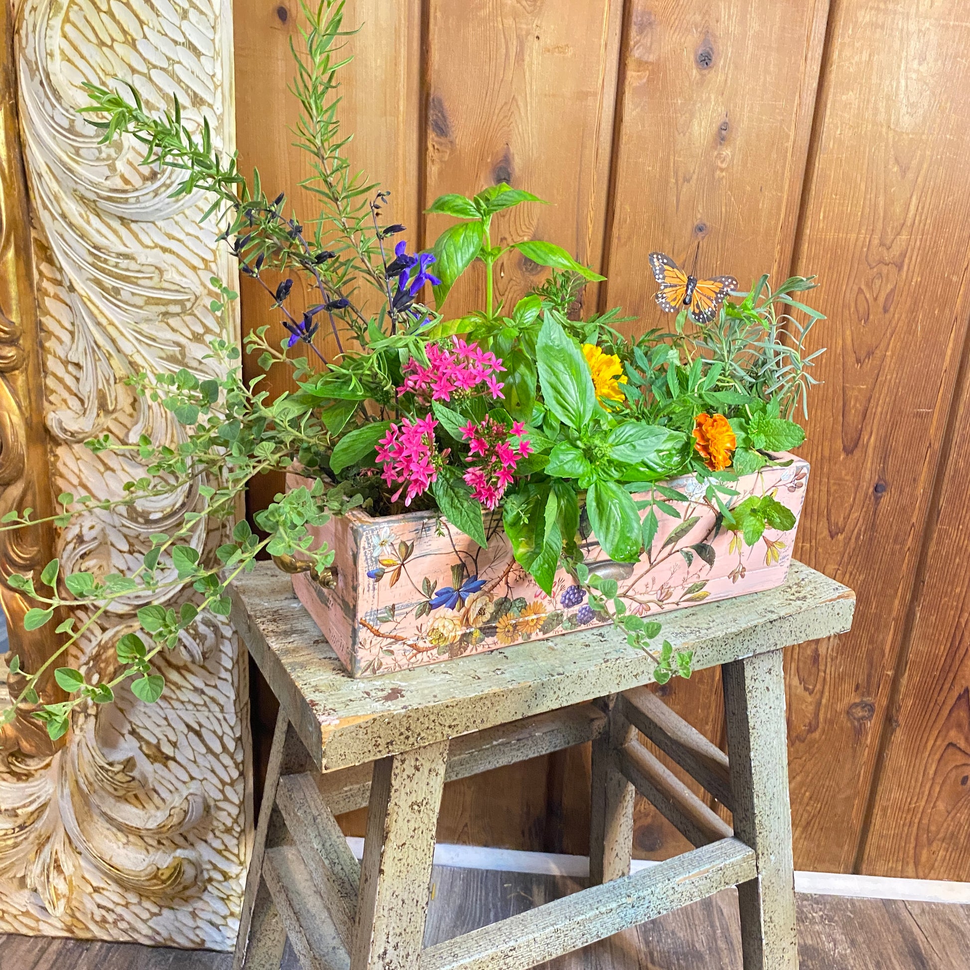 Decorative planter with flowers and herbs on a wooden stool against a wooden wall.