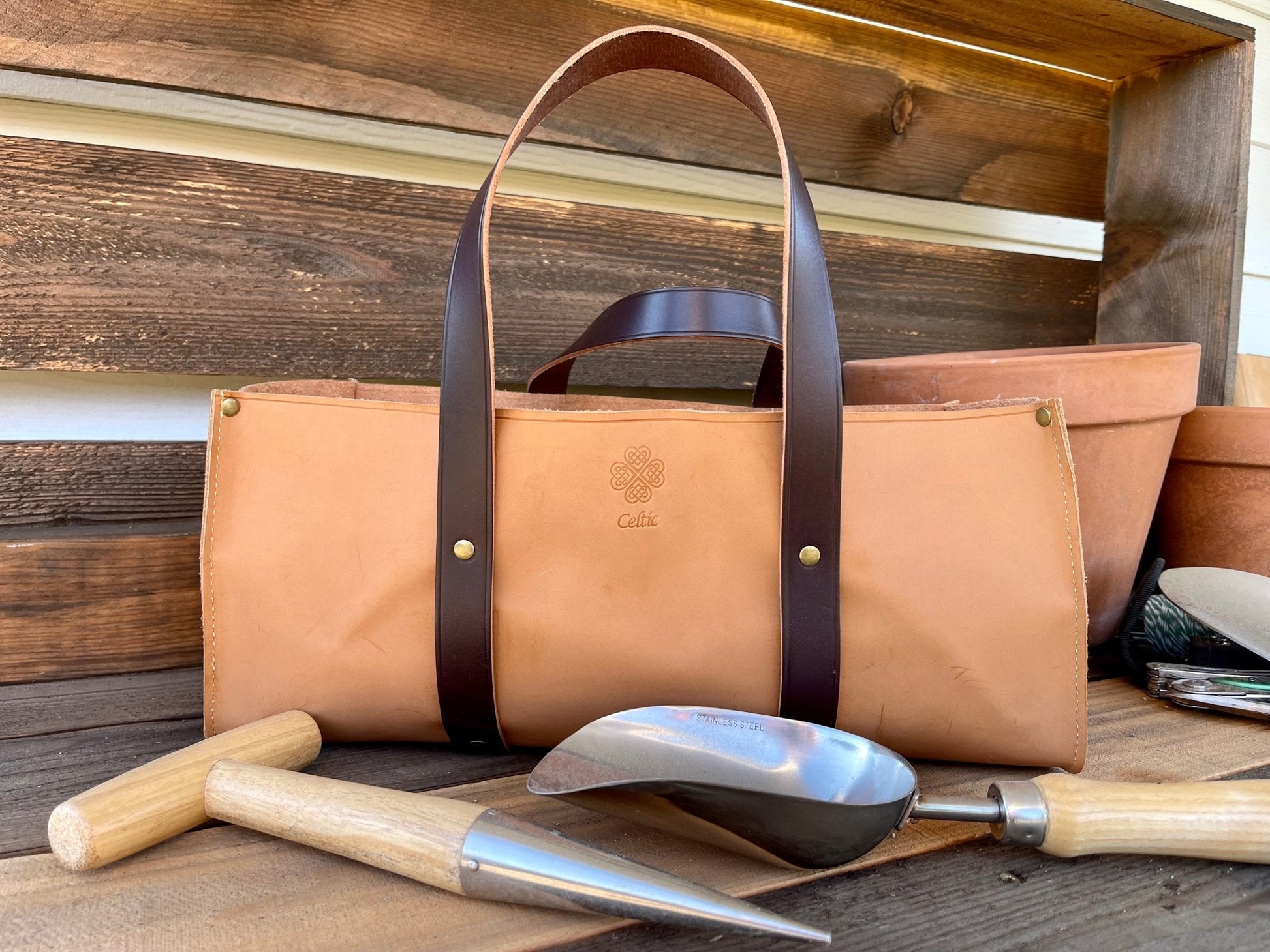 Brown leather tote bag with handles on a wooden surface with gardening tools.