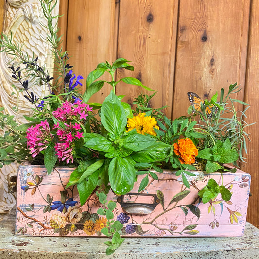 Decorative box with floral design filled with greenery and flowers against a wooden background